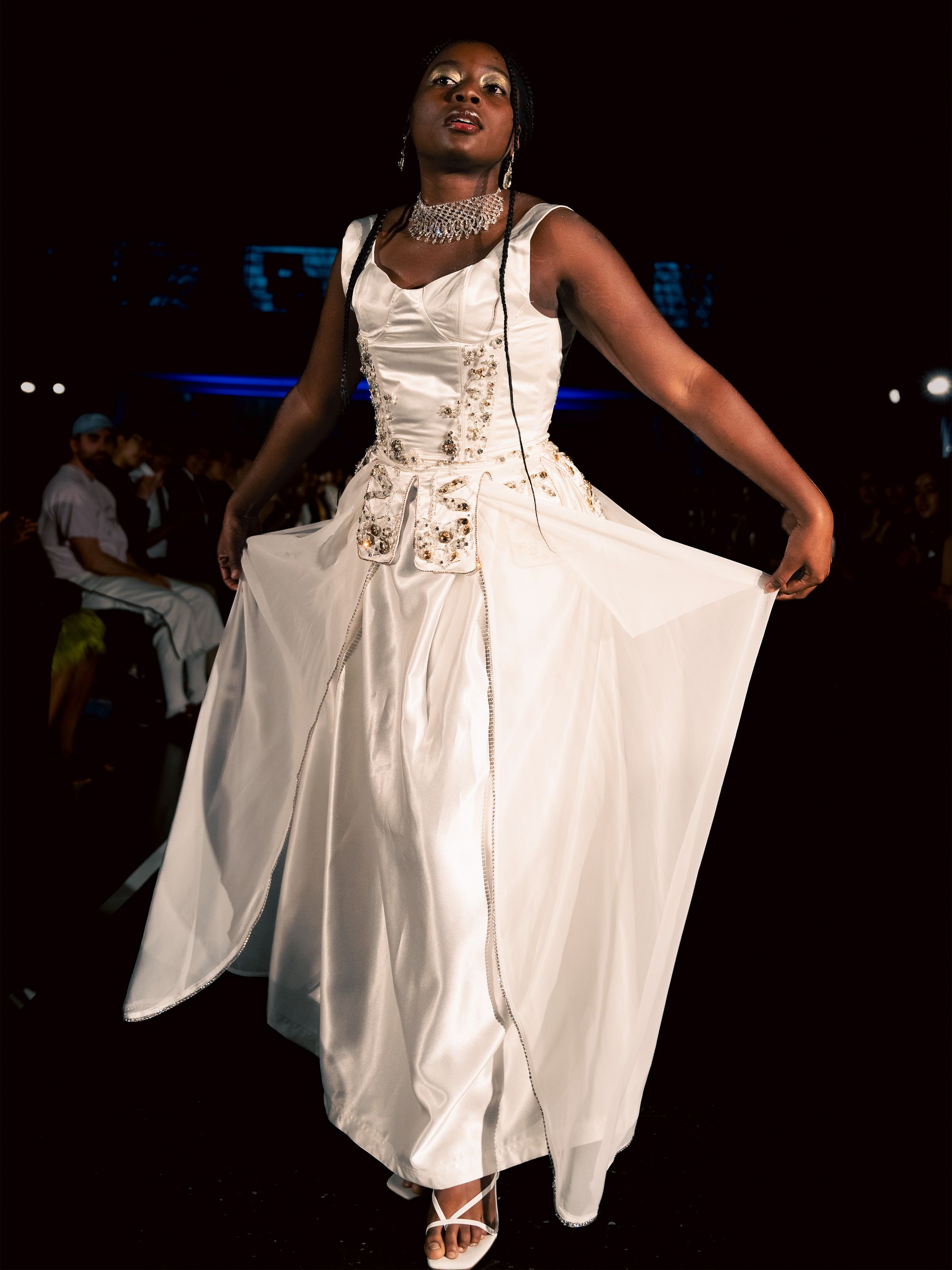 Model presents a white satin gown with a structured bodice and sparkling embellishments. The dress features layered sheer panels and is accessorized with a choker and white heels.